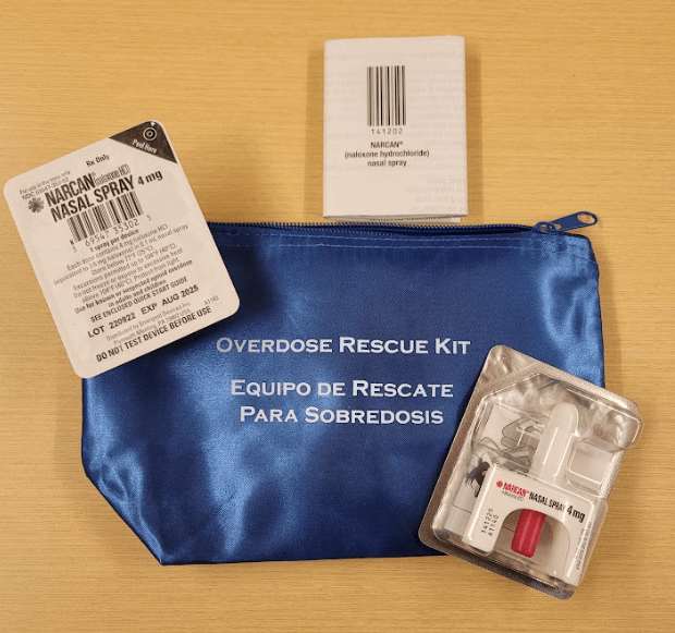 A blue, zipped pouch. Outside of the pouch are small plastic containers with Narcan Nasal spray inside. An instruction booklet is above them.