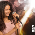 A young female person of color stands at a microphone with spotlights in the background.