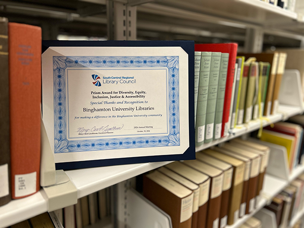 The SCRLC Prism Award for Diversity, Equity, Inclusion, Justice and Accessibility certificate is displayed on a Binghamton University Libraries collections shelf.