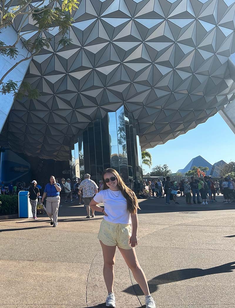 The geodesic sphere behind Maddie Clark houses the Spaceship Earth attraction where she is a cast member. This sphere has served as the symbolic landmark of EPCOT (Experimental Prototype Community of Tomorrow) since the park opened in 1982.