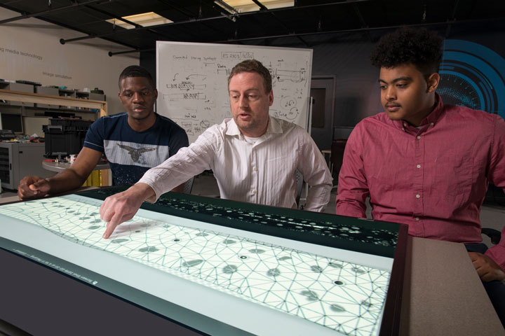 To straighten the image of a salamander, Associate Professor Scott Craver explains to Jonathan Milongo Kazadi, left, and Christian Tejera, right, how to draw a curved line that connects the points that have been plotted along the salamander's spine. They are working in the Seymour Kunis Media Core.